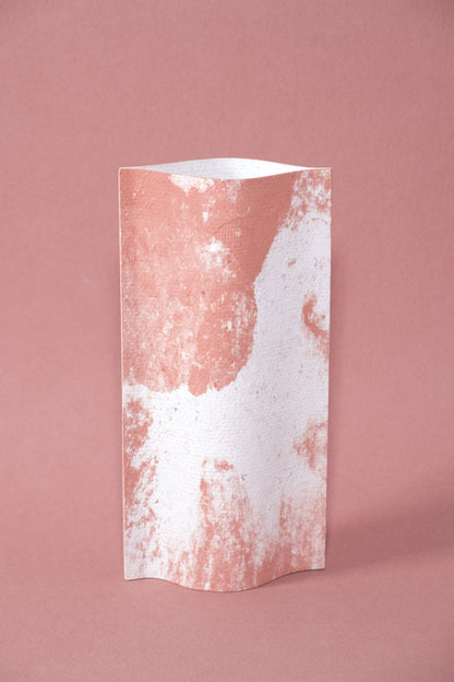 Un vase de profil rectangulaire sur fond rose. Deux feuilles de carton-pierre sont collées à leur extrémités et ouvertes en leur centre pour y laisser passer un contenant étanche.  Le vase arbore un effet marbré avec des veines couleur terra cotta sur fond blanc.