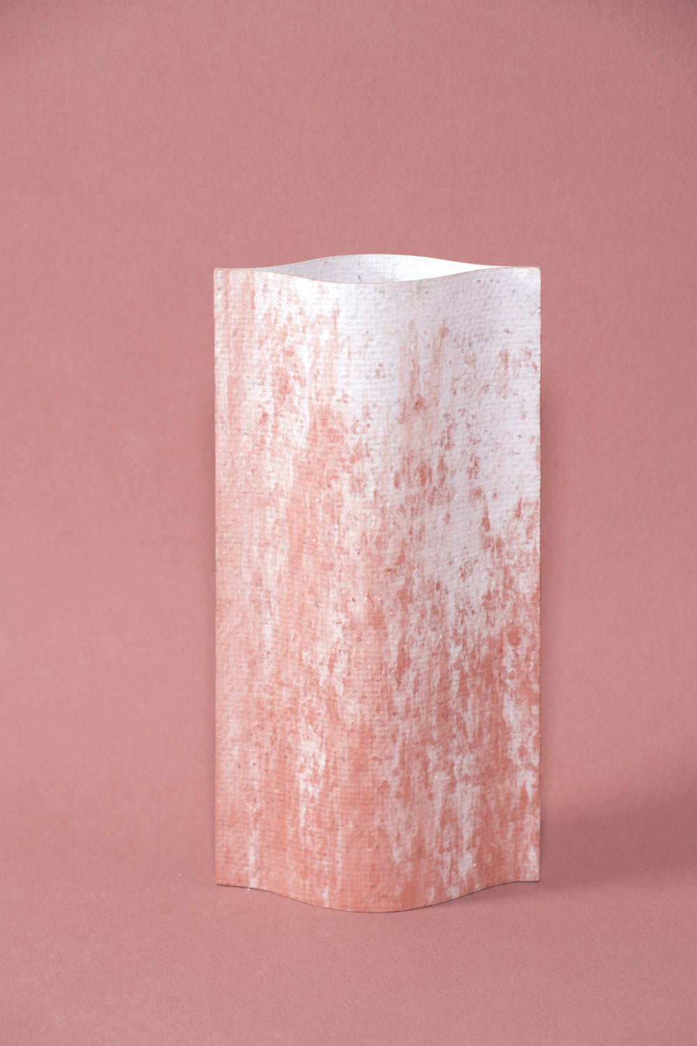 Un vase de profil rectangulaire sur fond rose. Deux feuilles de carton-pierre sont collées à leurs extrémités et ouvertes en leur centre pour y laisser passer un contenant étanche. Le vase arbore un effet marbré avec des veines couleur terra cotta sur fond blanc. Chaque vase a un marbré unique.