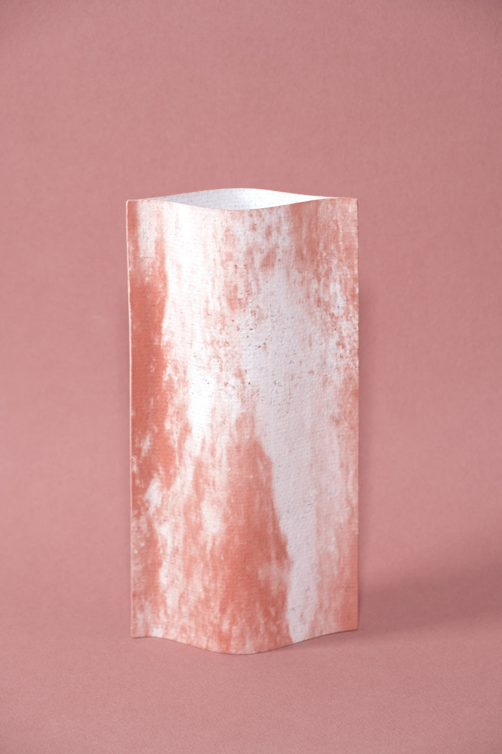 Un vase de profil rectangulaire sur fond rose. Deux feuilles de carton-pierre sont collées à leur extrémités et ouvertes en leur centre pour y laisser passer un contenant étanche.  Le vase arbore un effet marbré avec des veines couleur terra cotta sur fond blanc. Chaque vase a un marbré unique.