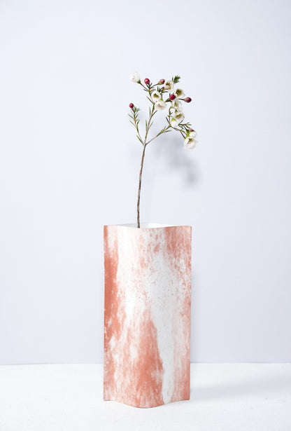 Un vase de profil rectangulaire sur fond blanc. Une branche y est soigneusement disposée. Deux feuilles de carton-pierre sont collées à leurs extrémités et ouvertes en leur centre pour y laisser passer un contenant étanche. Le vase arbore un effet marbré avec des veines couleur terra cotta sur fond blanc. Chaque vase a un marbré unique.