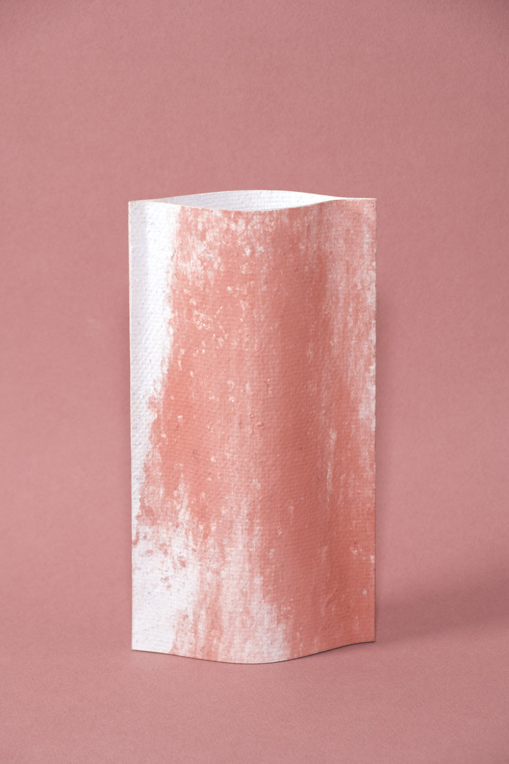 Un vase de profil rectangulaire sur fond rose. Deux feuilles de carton-pierre sont collées à leur extrémités et ouvertes en leur centre pour y laisser passer un contenant étanche.  Le vase arbore un effet marbré avec des veines couleur terra cotta sur fond blanc. Chaque vase a un marbré unique.