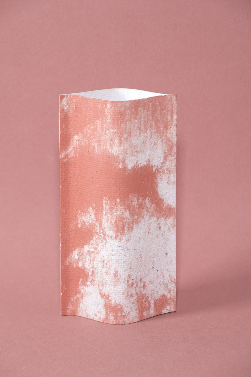 Un vase de profil rectangulaire sur fond rose. Deux feuilles de carton-pierre sont collées à leur extrémités et ouvertes en leur centre pour y laisser passer un contenant étanche.  Le vase arbore un effet marbré avec des veines couleur terra cotta sur fond blanc. Chaque vase a un marbré unique.