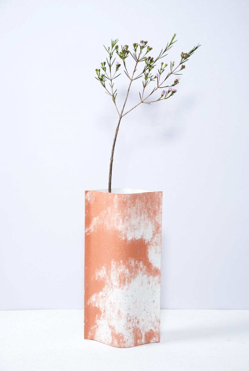 Un vase de profil rectangulaire sur fond blanc. Une branche y est soigneusement disposée. Deux feuilles de carton-pierre sont collées à leurs extrémités et ouvertes en leur centre pour y laisser passer un contenant étanche. Le vase arbore un effet marbré avec des veines couleur terra cotta sur fond blanc. Chaque vase a un marbré unique.