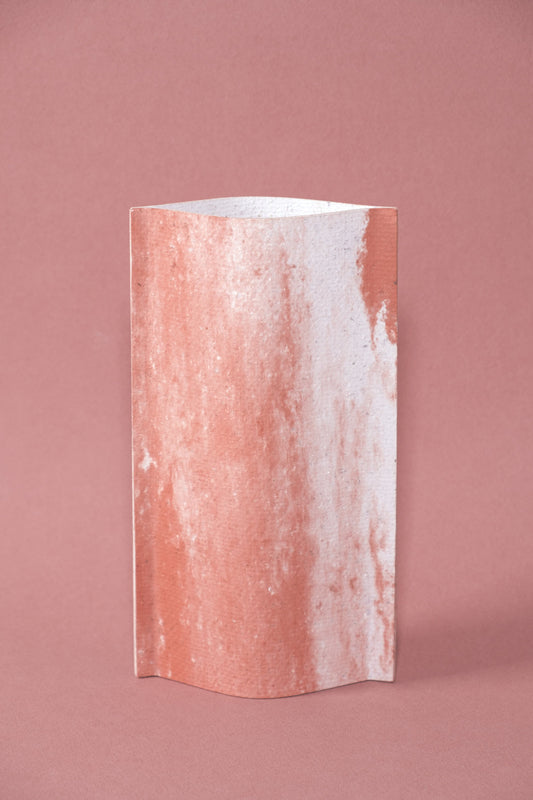 Un vase de profil rectangulaire sur fond rose. Deux feuilles de carton-pierre sont collées à leur extrémités et ouvertes en leur centre pour y laisser passer un contenant étanche.  Le vase arbore un effet marbré avec des veines couleur terra cotta sur fond blanc. Chaque vase a un marbré unique.