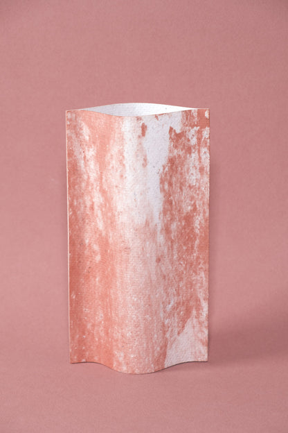 Un vase de profil rectangulaire sur fond rose. Deux feuilles de carton-pierre sont collées à leurs extrémités et ouvertes en leur centre pour y laisser passer un contenant étanche. Le vase arbore un effet marbré avec des veines couleur terra cotta sur fond blanc. Chaque vase a un marbré unique.