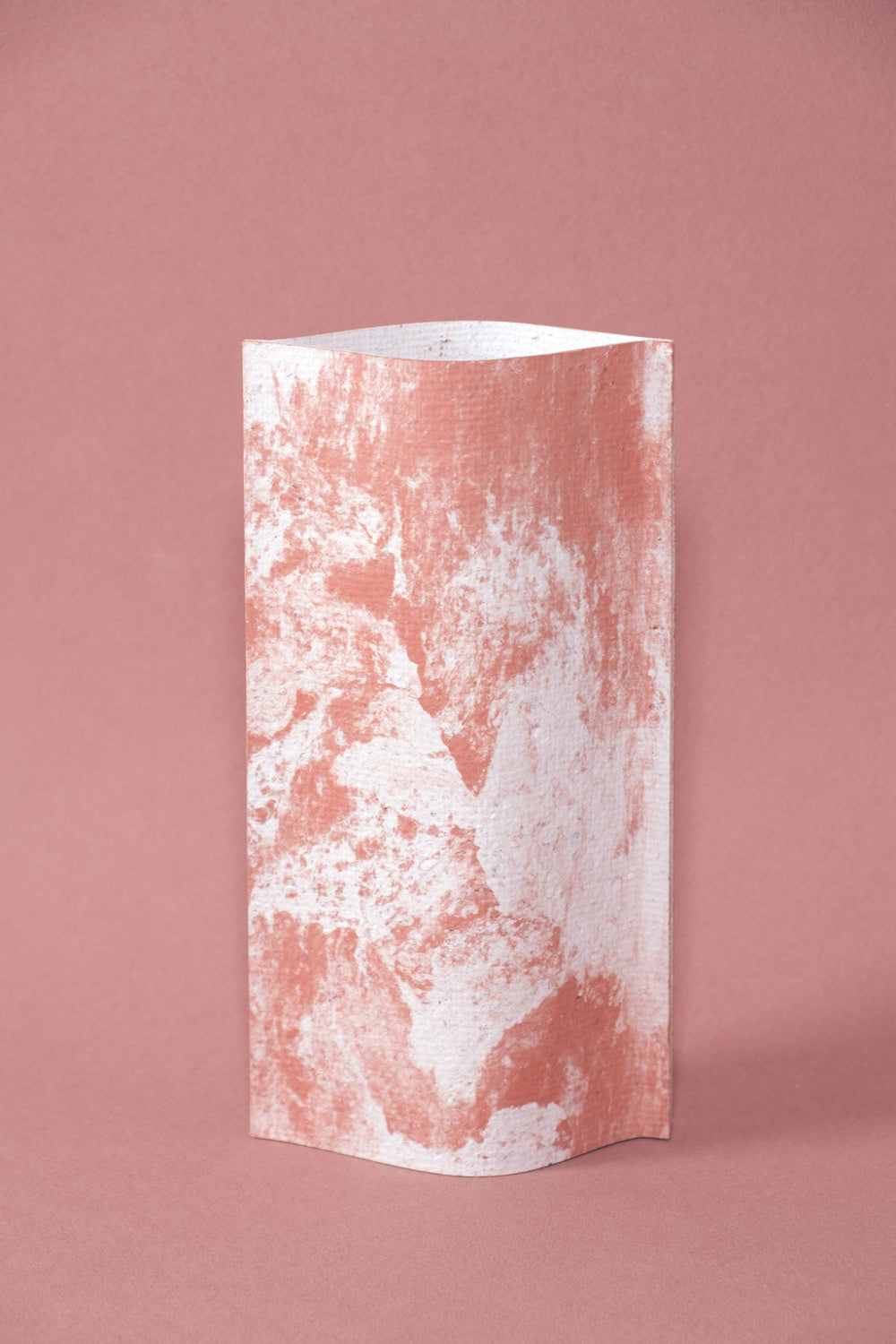 Un vase de profil rectangulaire sur fond rose. Deux feuilles de carton-pierre sont collées à leurs extrémités et ouvertes en leur centre pour y laisser passer un contenant étanche. Le vase arbore un effet marbré avec des veines couleur terra cotta sur fond blanc. Chaque vase a un marbré unique.