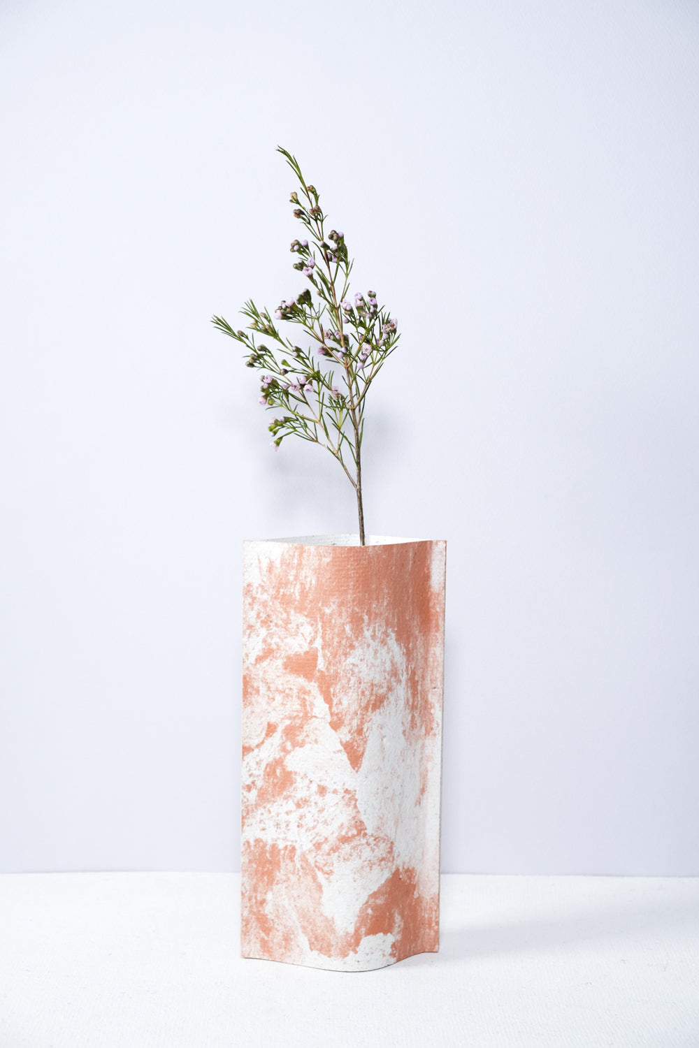 Un vase de profil rectangulaire sur fond blanc. Une branche y est soigneusement disposée. Deux feuilles de carton-pierre sont collées à leurs extrémités et ouvertes en leur centre pour y laisser passer un contenant étanche. Le vase arbore un effet marbré avec des veines couleur terra cotta sur fond blanc. Chaque vase a un marbré unique.