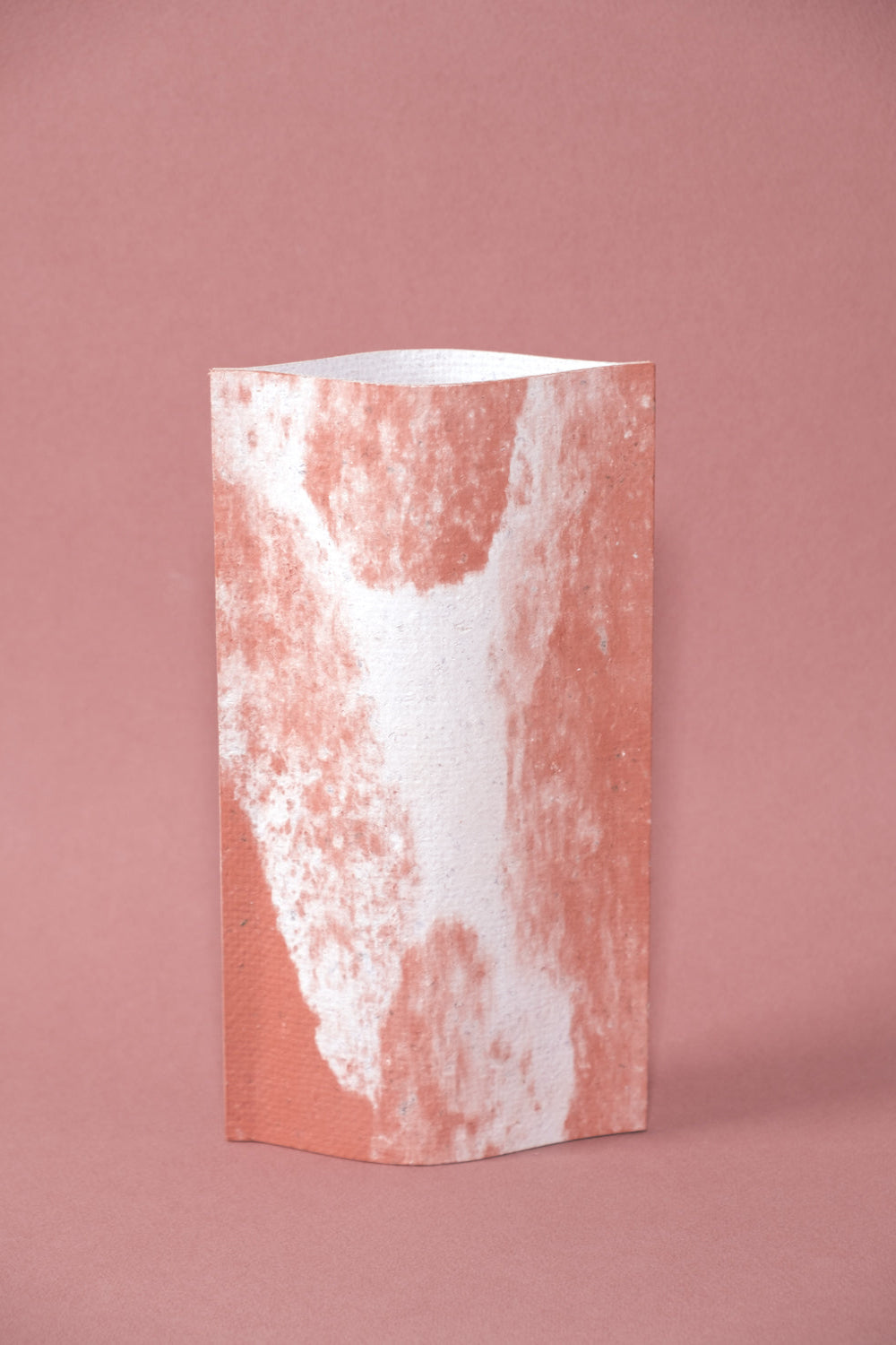 Un vase de profil rectangulaire sur fond rose. Deux feuilles de carton-pierre sont collées à leurs extrémités et ouvertes en leur centre pour y laisser passer un contenant étanche. Le vase arbore un effet marbré avec des veines couleur terra cotta sur fond blanc. Chaque vase a un marbré unique.