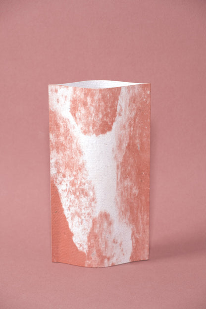 Un vase de profil rectangulaire sur fond rose. Deux feuilles de carton-pierre sont collées à leurs extrémités et ouvertes en leur centre pour y laisser passer un contenant étanche. Le vase arbore un effet marbré avec des veines couleur terra cotta sur fond blanc. Chaque vase a un marbré unique.