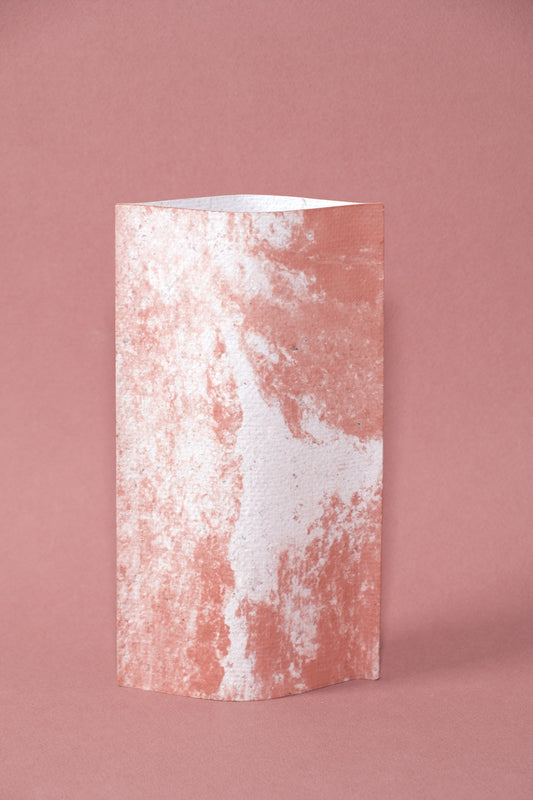 Un vase de profil rectangulaire sur fond rose. Deux feuilles de carton-pierre sont collées à leurs extrémités et ouvertes en leur centre pour y laisser passer un contenant étanche. Le vase arbore un effet marbré avec des veines couleur terra cotta sur fond blanc. Chaque vase a un marbré unique.