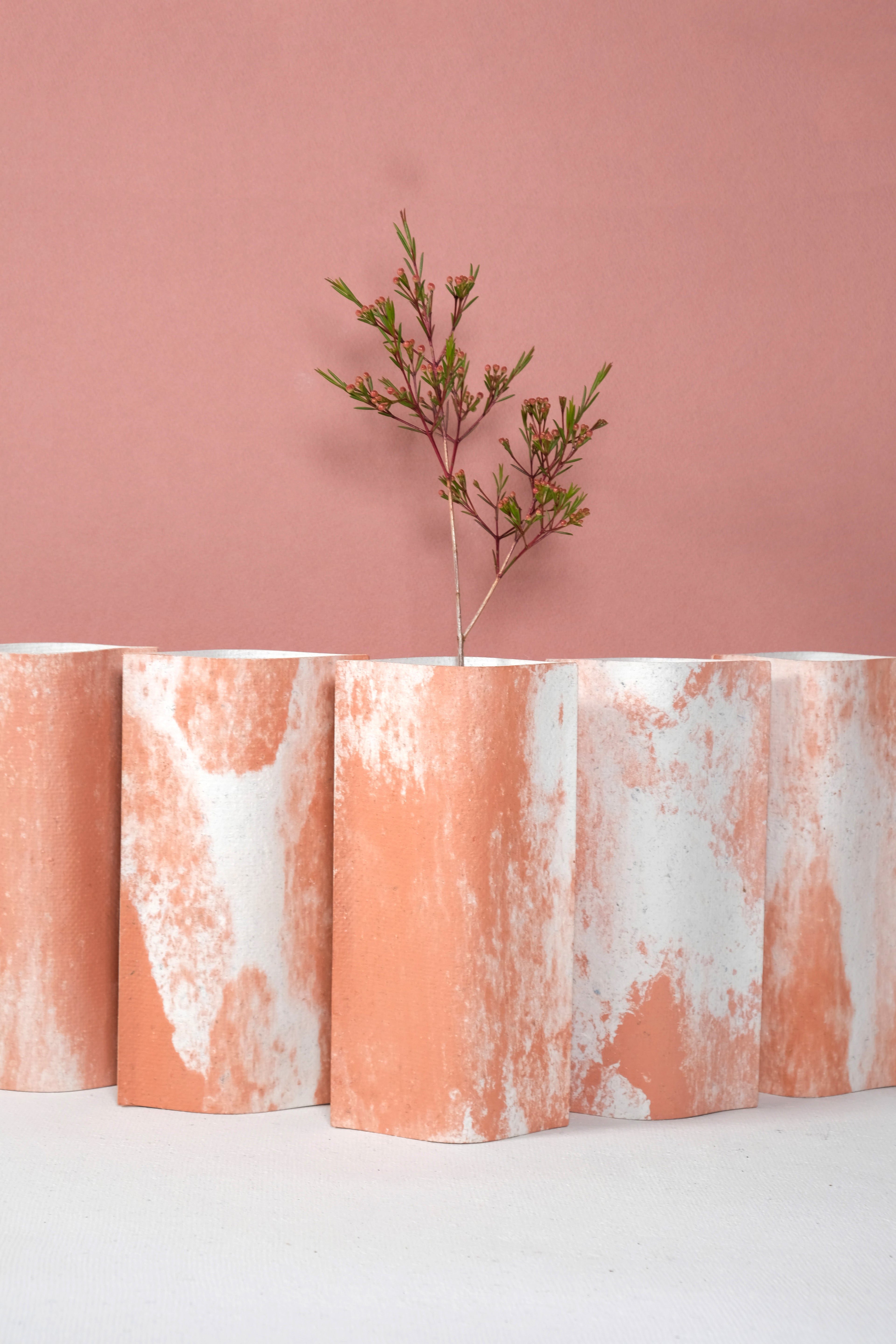 Ensemble de vases de la série terra cotta de profil rectangulaire sur fond rose. Ces vases sont fabriqués à partir de deux feuilles de carton-pierre collées à leurs extrémités et ouvertes en leur centre pour y laisser passer un contenant étanche. Les vases arborent un effet marbré avec des veines couleur terra cotta sur fond blanc. Chaque vase a un marbré unique.